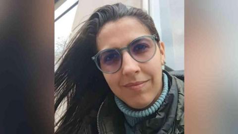 Vitória is looking at the camera up close. She is wearing a blue turtle neck jumper, a khaki or dark-coloured coat, has glasses on, and has brown hair.