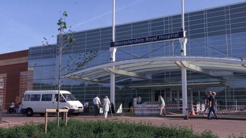 The exterior of Worcestershire Royal Hospital. It is a large glass-fronted building with a covered entrance. A white van is outside the building and a number of people are walking nearby.