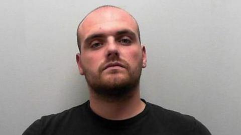 A police mugshot of a bald man, wearing a black shirt. He has light stubble and is staring at the camera.