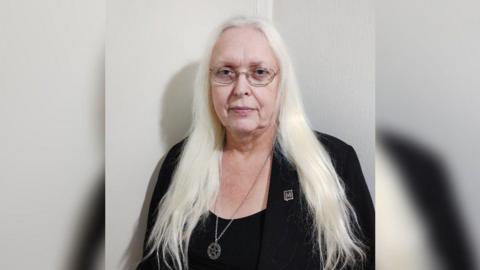 A formal mid-shot of Maz, who has long white hair and glasses, and is wearing a black vest and jacket