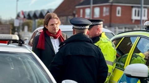 Police officers stand beside marked patrol cars, speaking with a man - Steff Aquarone - wearing a dark coat and red scarf. The scene takes place on a residential street with brick houses in the background.