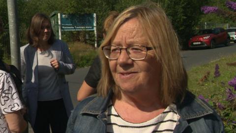 Linda Scott, who has shoulder length light brown hair and glasses, is wearing a denim jacket. She is standing with a group of other women near to the entrance to the site. 