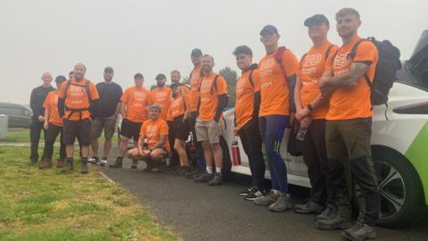 A group of 16 men and women stand together for a photo at the start of a 120km hike from Filey to Hull. They are wearing matching orange T-shirts and the weather is very grey and misty.