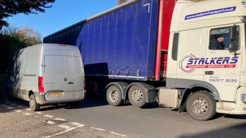 A grey van struggles to pass a blue lorry as it drives through a narrow road. 