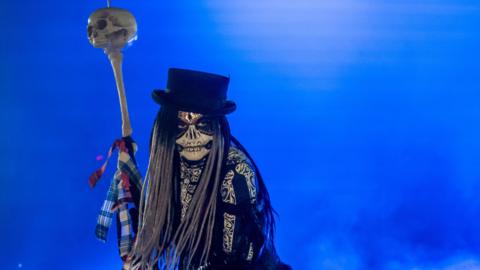 Shows a skeletal figure in a black top hat and a staff with a skull