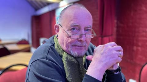 Max Bromley, a white, middle-aged man, photographed sitting on chair in front of a red curtain, wearing a green scarf and navy hoody with glasses on the end of his nose. He is sat with his elbows leaning on a table in front of him with his hands together in front of him. 