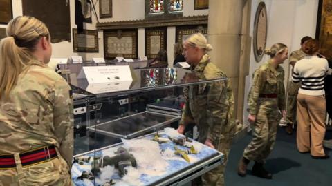 Two servicewomen in their camo uniform carrying a glass box containing a collection of knitted planes. People are doing similar behind them.