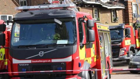 Red and yellow fire engine with "Bucksfire.gov.uk" in white lettering on the front. There is another fire engine just visible to the left and a third behind. A two-storey building with a collapsed fire-damaged roof is visible behind.