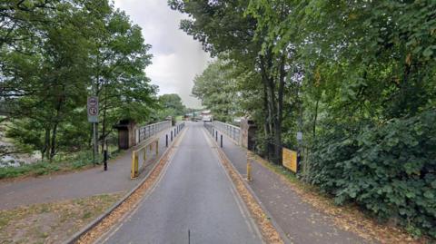 A view along Sheepmount Bridge. There is a single lane road across the bridge, which is lined with blue bollards. There are grey barriers running along the bridge. Trees can be seen on both sides of the river.