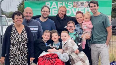 A photo of a group of men and women, with including four children and a baby. They all have their aims around each other and are smiling. Behind them is a banner saying '"Rafi's recovery".