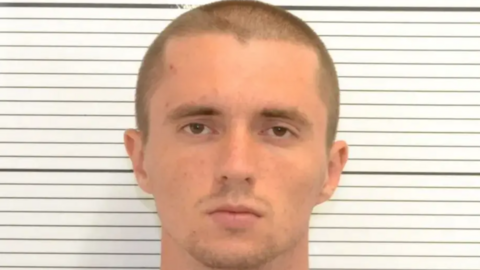 A white man with very short hair and stubble on his face stands in front of a black and white background for a police mugshot.