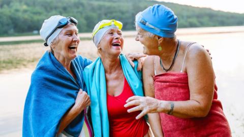 Three women in colourful swim caps and towels on the beach.