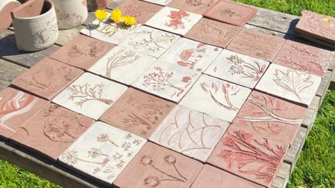 A range of tiles with different designs including plants, flowers and houses can be seen on a table. Three dandelions can be seen on the left. The table is on grass.