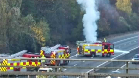 A traffic camera image above the M11 carriageway. Three fire engines block the road. Smoke from a car on fire can be seen billowing upwards from behind one of the fire engines.