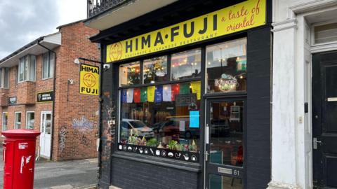 A Bhutanese and Japanese restaurant with a black exterior and a bright yellow sign reading “HIMA FUJI,” decorated with flags, plants and a neon “KARAOKE NIGHT” sign.