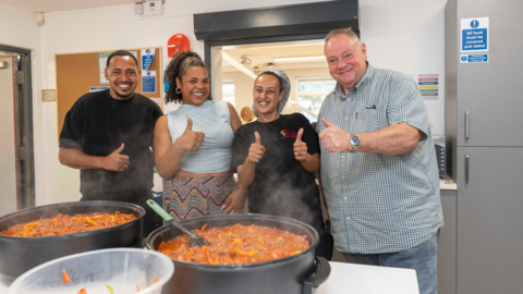 Three men and a woman stand behind two big cooking pots of food on a table in a kitchen. All four of them have their thumbs up and are smiling at the camera. There is a a large kitchen hatch behind them
