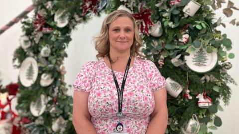 A woman with wavy hair in a pink floral top is looking at the camera. Behind her is a festive display including plates with Christmas trees on