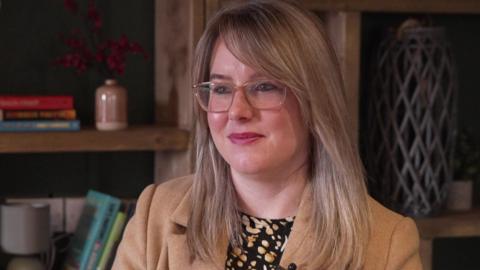Kirsty Walsh - The image shows a woman seated indoors against a backdrop of wooden shelving units. The shelves are filled with a mix of books, decorative items, and small ornaments. Some of the items include stacked books, a ceramic jug, a small vase, and a tall lattice-style decorative object. 
The woman is wearing a light brown or camel-coloured coat over a patterned top featuring black and pale shapes. 
