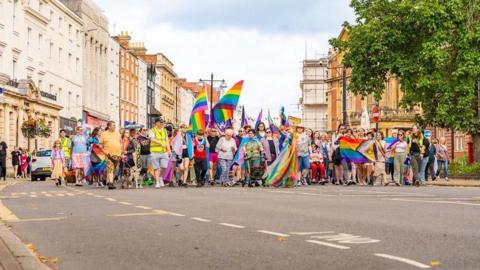 The start of the Pride march in Leamington with many people holding rainbow banners between the Georgian buildings of the town
