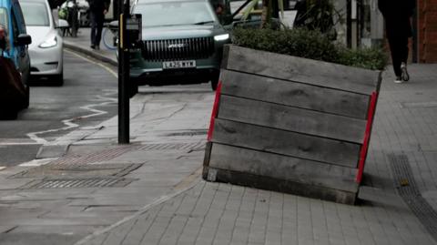 Pavement with a big planter on a part of the pavement sloped downwards. The background includes cars and pedestrians.