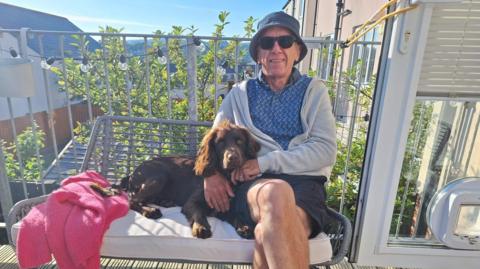 Gwyn Morley sat on a bench with a brown dog resting on his lap. Mr Morley has a navy blue sun hat on and sunglasses. He is wearing a blue patterned shirt, grey cardigan and navy blue shorts.