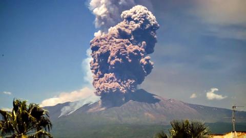 A view from the distance of Mount Etna as it erupts. A large smoke cloud billows into the sky.