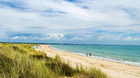 A sunny day at a beach. There is blue sky and golden sand with two people walking along the shoreline. You can see colourful windbreaks/tents in the distance and the beach has a strip of green dunes behind. 