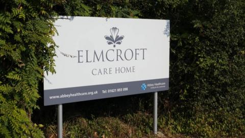 A grey and white sign that reads Elmcroft Care Home. It is surrounded by greenery.