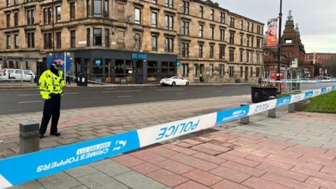 Police tape covering a crime scene where four people were arrested for serious assault. A policeman is standing behind it, with the road behind him.