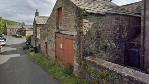 A old-looking stone built stable, set over two floors, flanked by a wall and another building. The property has a think strip of grass in front and then is directly onto a road.