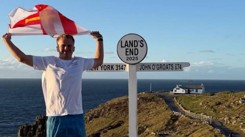Drew Jonkmans holds a Guernsey flag above his head at the Land's End road sign. He is wearing a white t-shirt and blue shorts. A building is at the end of a road in background. The sea is behind Drew. It is a sunny day with a few clouds in the sky.