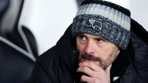 Paul Warne with his hand on his chin, wearing a Derby woolly hat