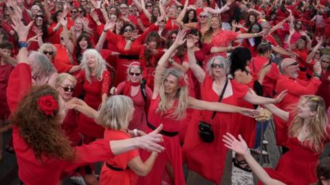 A group of people in red dresses dance with their arms in the air.