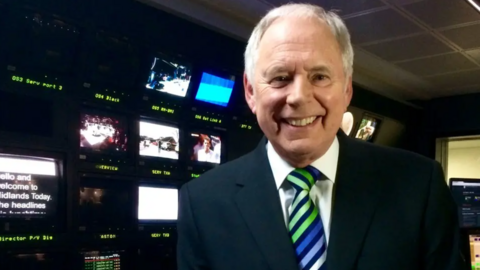 Nick Owen in a television studio. He has grey hair, is smiling and wearing a smart suit with a green, blue, silver, blakc striped tie. There are a number of television screens behind him and an autocue which readys hello and welcome to Midlands Today.