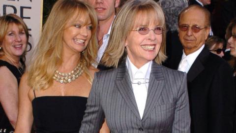 A smiling Diane Keaton dressed in a grey pinstripe suit and wearing glasses stands in front of Goldie Hawn, who is wearing a little black dress