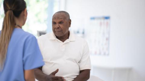 An older black men wearing a white shirt sits on a bed in a clinic talking to a nurse dressed in a blue uniform.