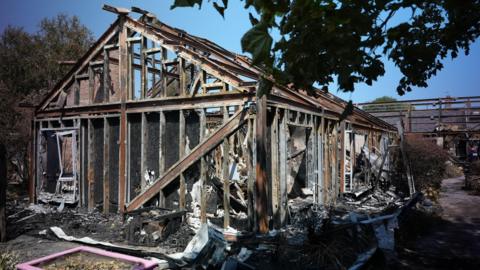 The charred remains of a collapsed hospital building stand amid scattered debris, with blackened wooden beams exposed.
