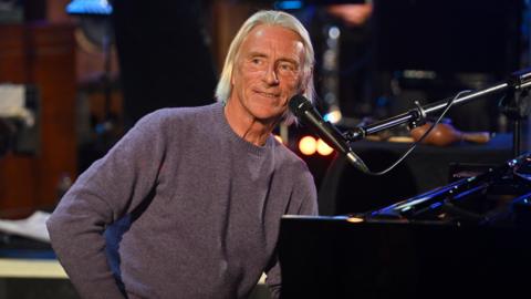 Paul Weller is sat at a piano, smiling. He has chin length blond hair and is wearing a purple jumper. 