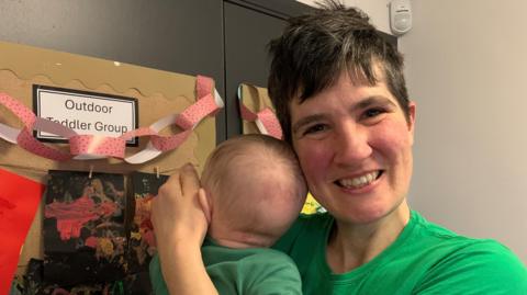 A woman with short black hair and wearing a bright green t-shirt is smiling. She is holding a baby to her shoulder, who is facing away. They are in front of a wall with information about toddler groups