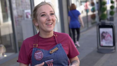 A 32 year old woman in a dark red costa uniform. She wears an apron reading barista and smiles at the camera. She has blonde hair pulled off of her face and stands in Bridgend town centre.