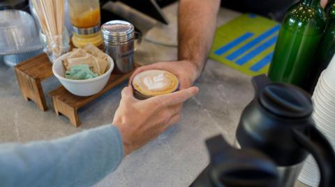 an arm in a blue sweater accepts a cappuccino with a leaf decal in the foamed milk 