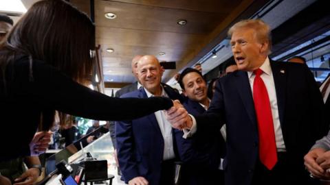 Donald Trump in Dearborn, Michigan wearing a blue coat and tie. He is smailing and reaching forward to shake hands with a woman whose face we can't see in a cramped space surrounded by people.