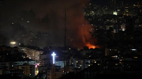 Smoke and orange flames rise, following an Israeli strike on Beirut's southern suburbs. A glowing white cross chan be seen in the foreground 