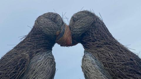 A side on shot of the two puffin sculptures with their heads together. The willow is frayed and coming off in many places and the paintwork is faded. A grey-ish sky is visible in the background.