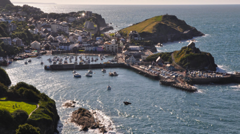 The picture shows a coastal harbour surrounded by steep green hills and a cluster of buildings stretching up the slopes. The water is a deep blue and several small boats are moving in and out of the sheltered harbour area. There are piers curving around the water to protect the boats, and rocky outcrops sit at the harbour entrance where the sea meets the land.