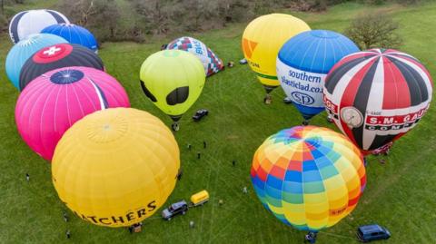 12 hot air balloons are shown blown up in a green field. There are some vans and people dotted around.