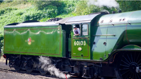 A green steam train runs through the countryside while the driver in a white shirt, dark-coloured apron and cap, leans out of his window.