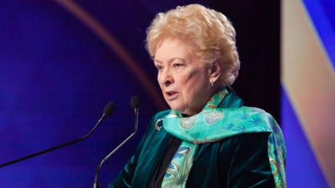 Baroness O'Loan in green suit and scarf giving a speech at microphone