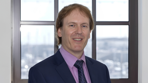 Richard Hemans wearing a blue suit and pink shirt. He has short brown hair and is stood in front of a window. 