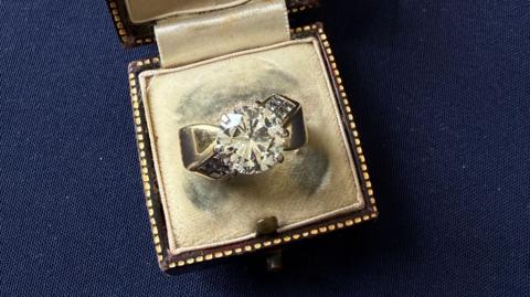 A diamond ring in an open vintage brown ring box on a dark blue background